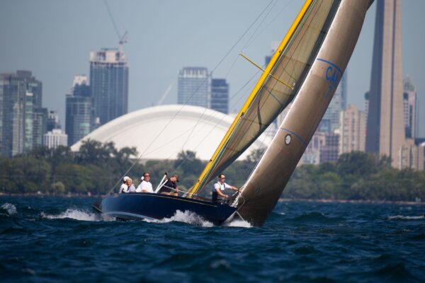 Sailing yacht International 8mR (1934) on water with city skyline backdrop.