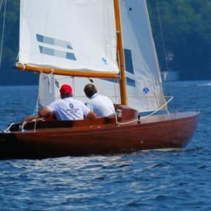 1957 Dragon 29 sailboat on a lake with two people, Canadian flag, and distant trees.